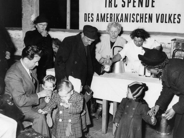 Menschen verteilen Lebensmittel an Menschen verschiedenen Alters. Im Hintergrund ist ein Plakat mit der Aufschrift: IRC Spende des amerikanischen Volkes.