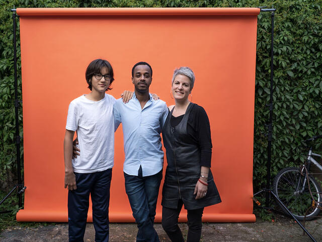 Ein Junge, ein junger Mann und iene Frau stehen vor einem orange farbenden Hintergrund und lächeln in die Kamera