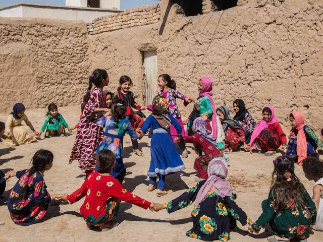Mädchen in bunter Kleidung sitzen oder stehen in einem Kreis und spielen fröhlich. Sie sind draußen auf einem trockenen Boden.