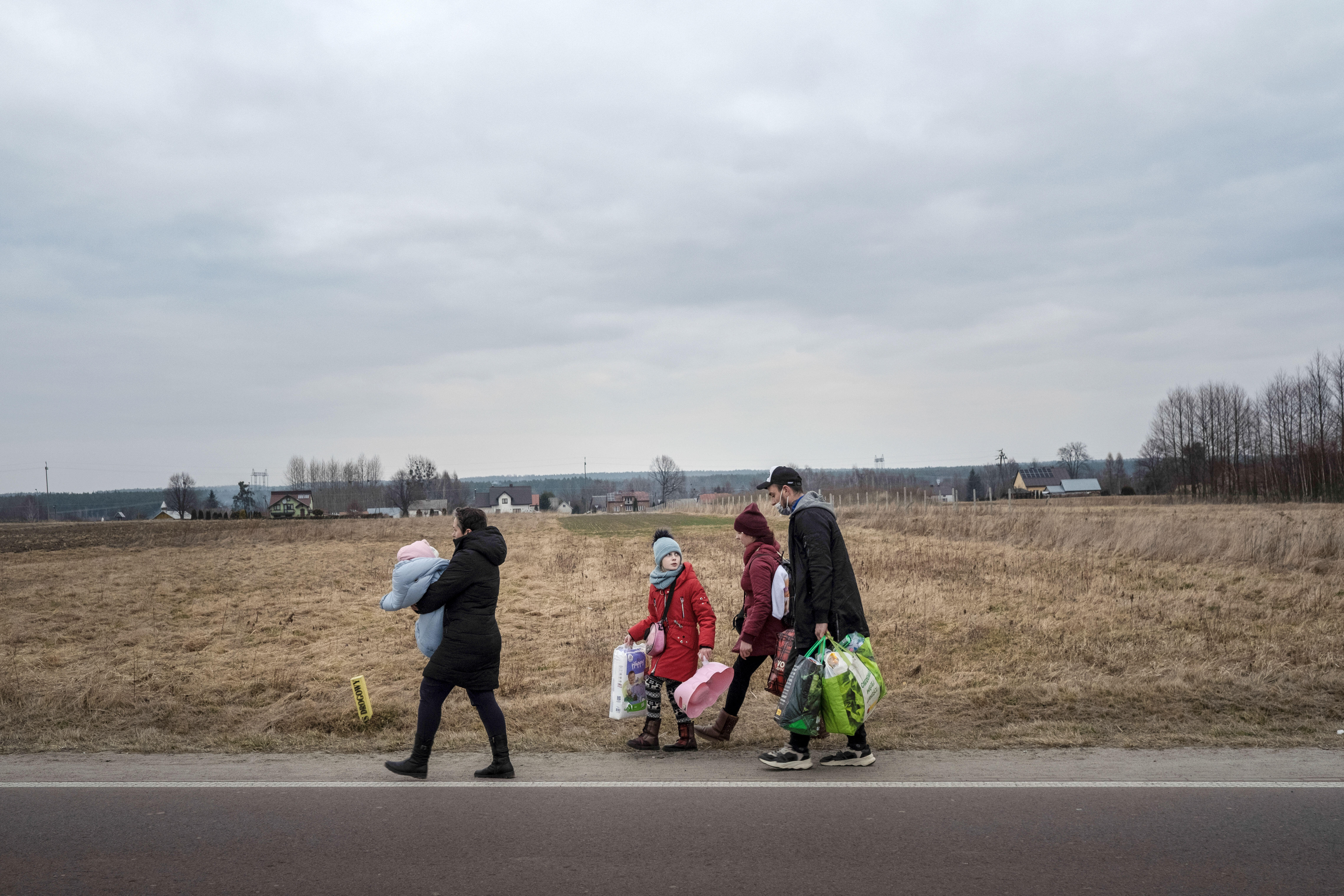 Eine Familie mit drei Kindern läuft zu Fuß mit vielen Taschen und Tüten eine Landstraße lang