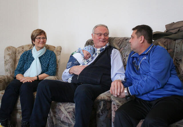 Eine Frau und zwei Männer sitzen auf einem Sofa, der Mann in der Mitte hält ein Baby im Arm