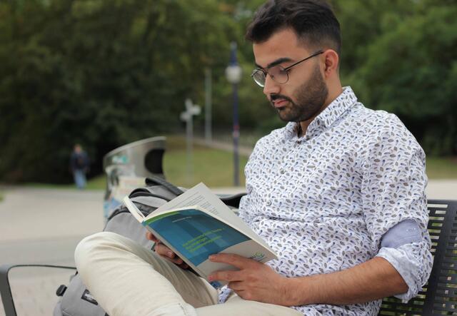 Zain sitzt auf einer Bank und liest in einem Buch 