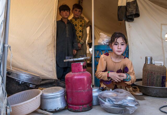 Ein Mädchen hockt mit Kochutensilien vor einem Zelt, zwei Brüder stehen im Hintergrund.