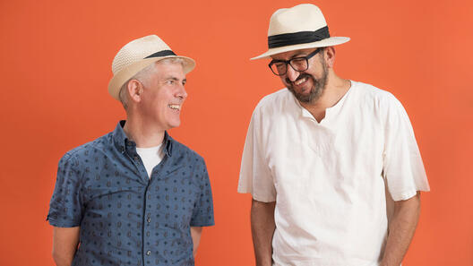 Zwei Männer, die beide Hüte tragen, stehen vor einer orangenen Wand und lachen.